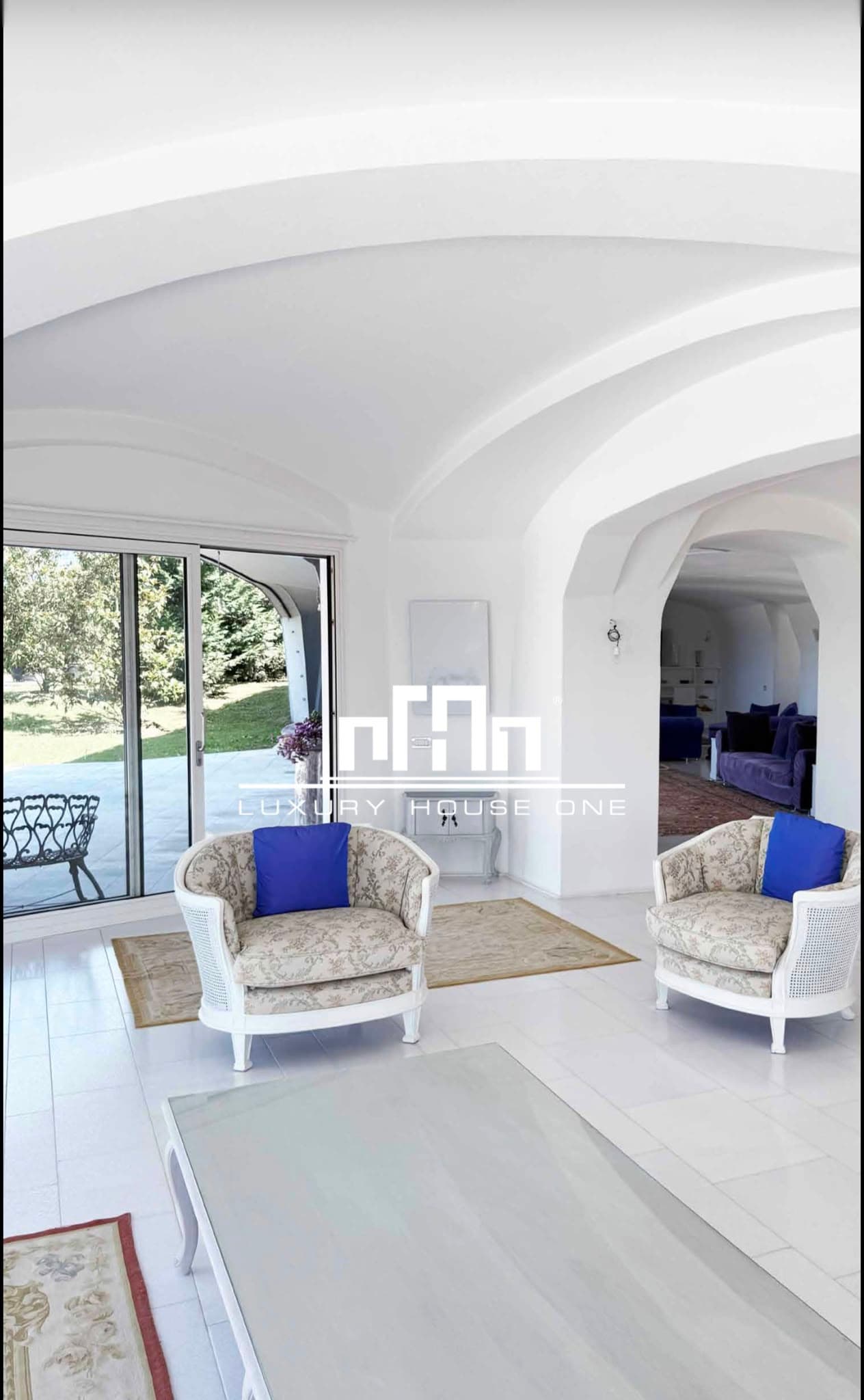 Soggiorno bianco con soffitti a volta, poltrone fiorite e vista panoramica sul giardino esterno.