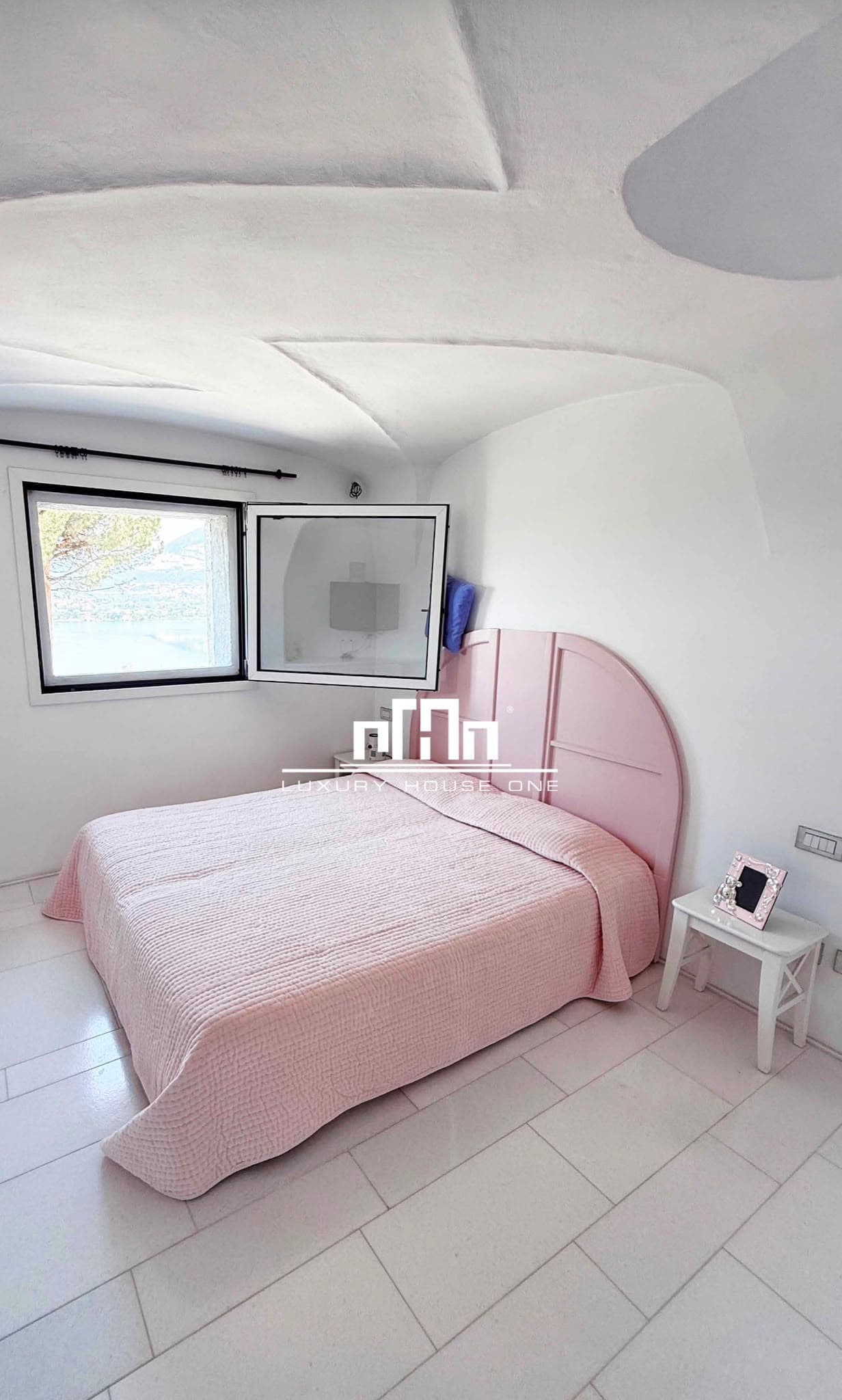 Camera bianca con soffitto a volta, letto rosa e finestra aperta con vista mare.